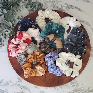 Może przedstawiać: Kolekcja 11 kolorowych scrunchie w różnych wzorach i kolorach, w tym zielonym, niebieskim, czarnym, białym i różowym. Scrunchie są ułożone na drewnianej powierzchni.