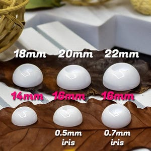 Może przedstawiać: Białe kaboszony w kształcie kopuły w różnych rozmiarach, w tym 18 mm, 20 mm i 22 mm. Mniejsze rozmiary są również pokazane, z wymiarami takimi jak 0,5 mm tęczówki i 0,7 mm tęczówki. Kaboszony są wyświetlane na brązowym liściu.