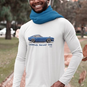 May include: White long-sleeve shirt featuring a blue sports car graphic with the text "Legends Never Die 240Z". The shirt is worn with a blue scarf and a gray beanie. The person is smiling.