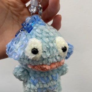 May include: A handmade blue and white crocheted creature keychain with black eyes and a peach-colored mouth. The keychain has a blue ring and is adorned with clear and blue beads and charms.
