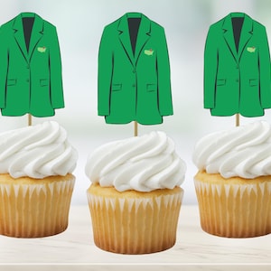 May include: Three cupcakes with white frosting, each topped with a green jacket-shaped pick. The jacket picks have a small logo on the left side. The cupcakes sit on a light-colored surface.