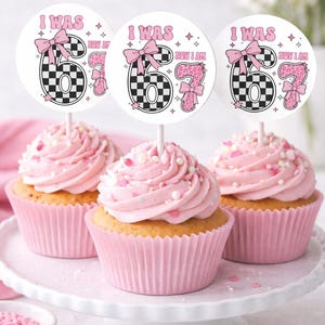 May include: Three cupcakes with pink frosting and sprinkles, each topped with a circular pick. The picks read "I WAS NOW I AM" with a checkered "6" and a pink "7" with a bow. The cupcakes sit on a white cake stand.
