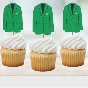 May include: Three cupcakes with white frosting, each topped with a green jacket-shaped decoration. The cupcakes sit on a light-colored surface. The jacket decorations have a black lapel and a small emblem.