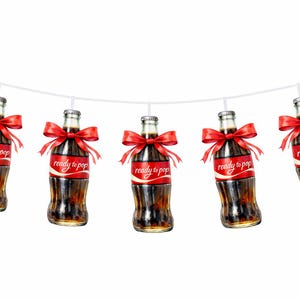 May include: A string of five soda bottle decorations. Each bottle has a red label with the text "ready to pop" and a red bow. The bottles are suspended from a white string against a white background.