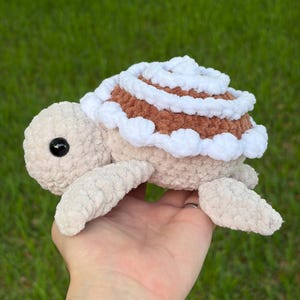 Peut inclure: Une tortue en crochet peluche avec un corps beige doux et une carapace ronde. La carapace présente un motif en spirale alternant du fil marron et blanc, avec un bord festonné blanc. La tortue possède un œil de sécurité noir visible.