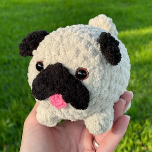 May include: A handmade, plush pug dog toy. The toy is cream-colored with black accents on the ears and snout. It has black eyes and a pink tongue. The toy is held in a hand against a green background.