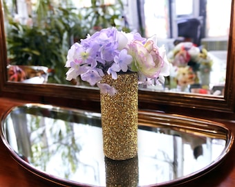 May include: A gold glitter vase holds a bouquet of artificial flowers. The flowers are a mix of purple, white, and pink. The vase sits on a reflective, oval-shaped tray. A mirror with a gold frame is in the background.