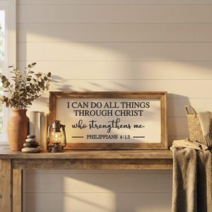 May include: A rectangular wooden sign with a white background and black text that reads I CAN DO ALL THINGS THROUGH CHRIST who strengthens me. PHILIPPIANS 4:13. The sign is framed in natural wood and sits on a wooden surface next to a lantern.