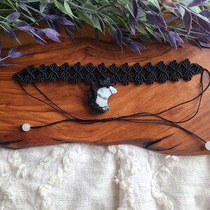 May include: Black macrame choker necklace with a white and green crescent moon pendant. The necklace is on a wooden surface with white fabric in the background.