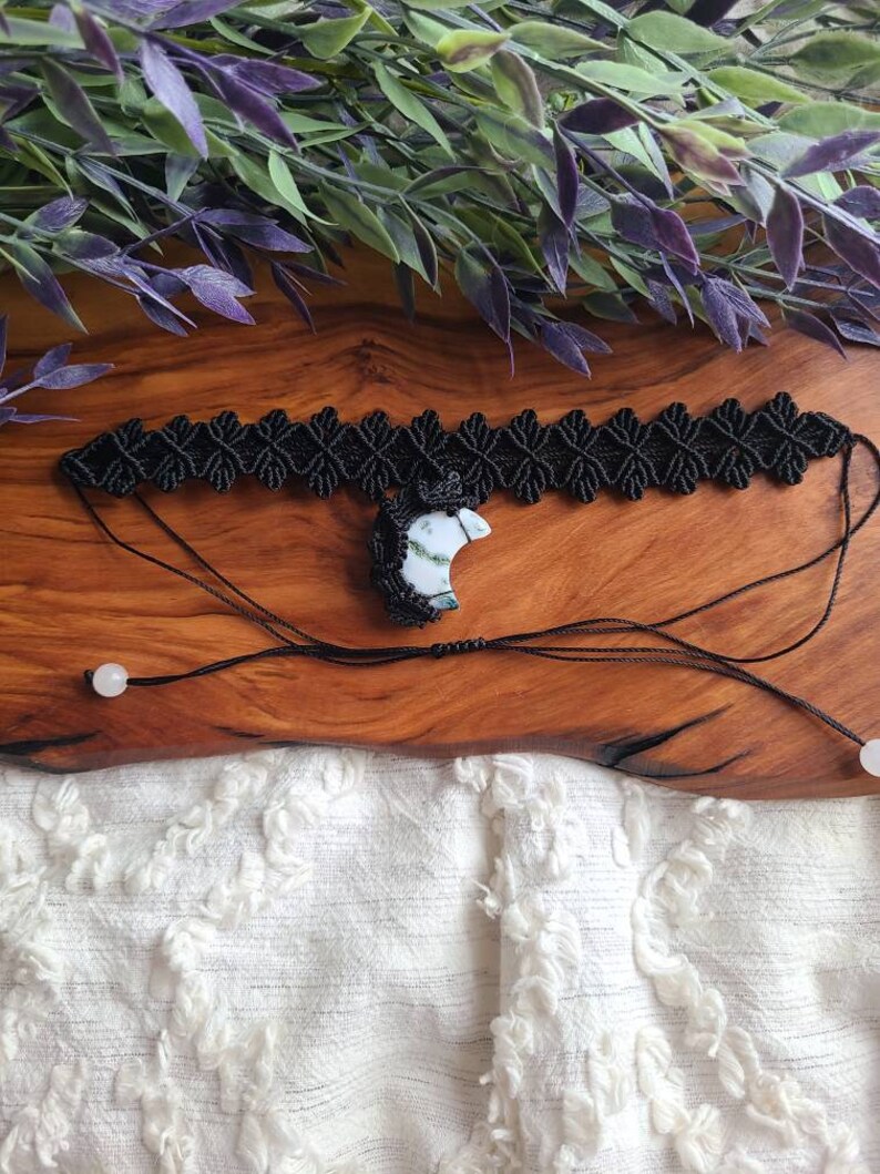 May include: Black macrame choker necklace with a white and green crescent moon pendant. The necklace is on a wooden surface with white fabric in the background.