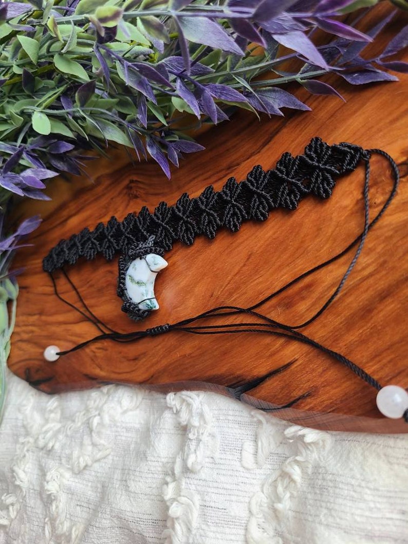 May include: A black macrame choker necklace with a white and green stone pendant. The necklace is tied with black cord and has white beads on the ends.