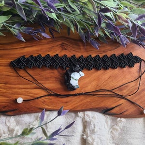 May include: Black macrame choker necklace with a white and green agate pendant. The necklace is on a wooden surface with green leaves in the background.