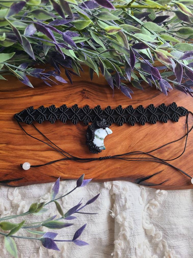 May include: Black macrame choker necklace with a white and green agate pendant. The necklace is on a wooden surface with green leaves in the background.