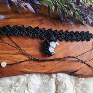 May include: A black macrame choker necklace with a white and green agate pendant. The necklace is on a wooden surface with green leaves in the background.