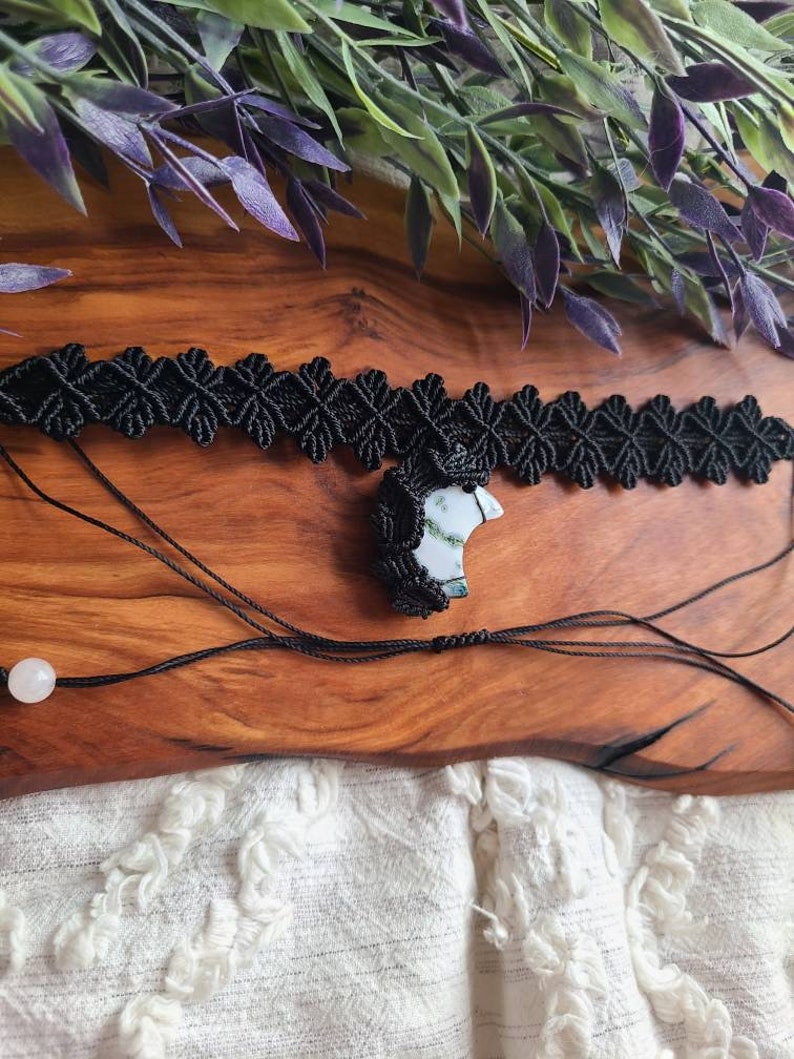 May include: A black macrame choker necklace with a white and green agate pendant. The necklace is on a wooden surface with green leaves in the background.