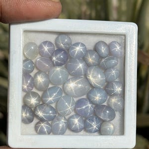 Pode incluir: Um close-up de uma coleção de safiras estrela azuis em uma caixa branca. As safiras são de forma oval e têm um padrão de estrela no centro.