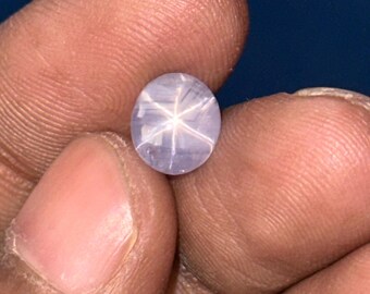 Könnte beinhalten: Ein runder, hellblauer Edelstein mit einem sechszackigen Sternmuster. Der Edelstein wird zwischen Fingern vor einem dunkelblauen Hintergrund gehalten. Der Sterneffekt entsteht durch Lichtreflexionen im Stein.
