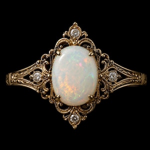 Könnte beinhalten: Ein goldener Ring mit einem ovalen Opal-Edelstein, umgeben von einem filigranen Design. Der Ring hat kleine Diamantakzente und ist vor einem schwarzen Hintergrund platziert. Die filigranen Details und warmen Farbtöne erzeugen eine elegante Ästhetik.