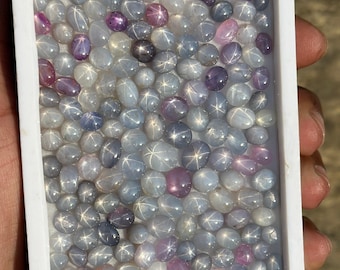 May include: A white tray filled with a variety of small, oval-shaped, star sapphire gemstones. The gemstones are a mix of light blue, gray, and pink.