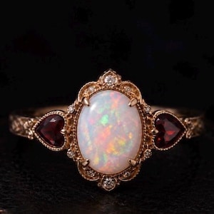 Può includere: Anello dorato con pietra centrale di opale ovale dai colori iridescenti. Due gemme rosse a forma di cuore affiancano l'opale. L'anello presenta intricati dettagli in filigrana e piccoli diamanti, su sfondo scuro.
