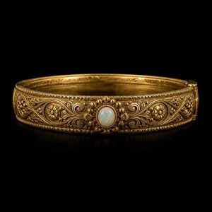 Puede incluir: Un brazalete dorado con intrincados diseños florales y de volutas. El brazalete presenta una piedra de ópalo ovalada en el centro. El metal tiene un acabado texturizado y antiguo, sobre un fondo negro.