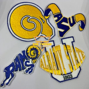 May include: Four embroidered patches featuring the logos of different organizations. The patches are white, blue, and yellow. The first patch is a yellow ram's head with a blue outline. The second patch is a blue and yellow ram's head with the word "RAMS" in blue. The third patch is a blue and yellow logo with the letters "ASU" in blue. The fourth patch is a white patch with the words "ALBANY STATE" in yellow and the year "1903" in blue.