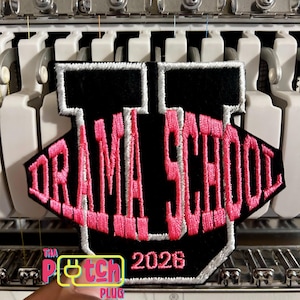 May include: A black and pink embroidered patch with the words "DRAMA SCHOOL" in pink, outlined in white, and the year "2026". The patch is on a white and black machine.