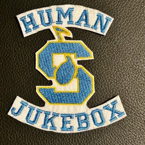 May include: A white fabric patch featuring the blue embroidered text HUMAN at the top and JUKEBOX at the bottom. In the center is a large blue letter S with a yellow outline and a small yellow musical note icon positioned above the letter.