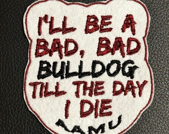 Può includere: Una toppa ricamata bianca con bordo rosso che presenta il testo I'LL BE A BAD, BAD BULLDOG TILL THE DAY I DIE in filo rosso e nero, con le lettere AAMU ricamate in nero nella parte inferiore su uno sfondo bianco simile al feltro.