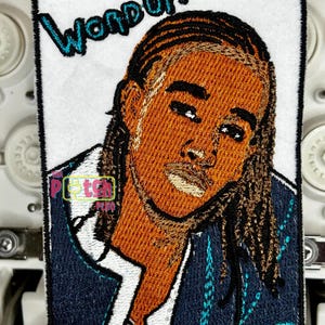 May include: Embroidered patch featuring a portrait of a person with long hair, wearing a dark blue jacket over a white shirt. The text "WORD UP!" is embroidered in teal above the portrait. The patch has a white background and a black border.