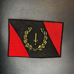 May include: A rectangular embroidered patch featuring a diagonal design with red and black sections. In the center, a yellow embroidered laurel wreath encircles a yellow vertical sword symbol, creating a detailed emblem on a fabric base.