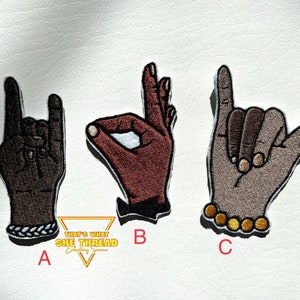 May include: Three embroidered iron-on patches depicting different hand gestures. The first patch shows a hand making the rock and roll sign, the second patch shows a hand making the ok sign, and the third patch shows a hand making the shaka sign. The patches are labeled A, B, and C.