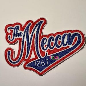 May include: Embroidered patch with the words "The Mecca" in a stylized script, with a red and blue color scheme. The year "1867" is below the text. The patch has a white border.