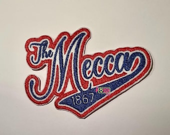 Può includere: Toppa ricamata con la scritta "The Mecca" in un carattere stilizzato, con una combinazione di colori rosso e blu. È incluso anche l'anno "1867". La toppa ha un bordo bianco.