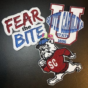 May include: Three embroidered patches featuring a white bulldog wearing a blue cap and red sweater with SC, a patch with the text FEAR the BITE in red and blue, and a patch with a large U, the text SOUTH CAROLINA STATE 1896, and a blue football.