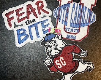 Può includere: Tre toppe ricamate raffiguranti un bulldog bianco con berretto blu e maglione rosso con SC, una toppa con il testo FEAR the BITE in rosso e blu, e una toppa con una grande U, il testo SOUTH CAROLINA STATE 1896 e un pallone da football blu.