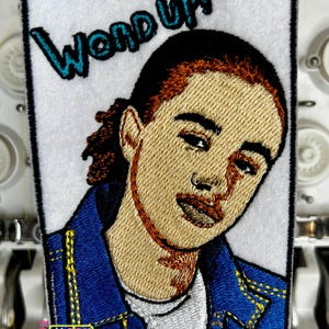 May include: Embroidered patch featuring a portrait of a person with the text "WOND UP!". The patch has a white background with a black border. The person is wearing a blue jacket with yellow stitching.