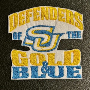 May include: A white fabric patch with embroidered text and a central logo. The text reads DEFENDERS OF THE GOLD & BLUE in yellow and blue thread. A stylized SJ logo is centered in blue and yellow. The patch has a white felt-like background.