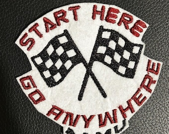 Può includere: Una toppa ricamata bianca con due bandiere da corsa a scacchi bianche e nere incrociate. Il testo "START HERE" è arcuato in rosso sopra le bandiere, "GO ANYWHERE" è arcuato in rosso sotto, con "AAMU" in nero nella parte inferiore.