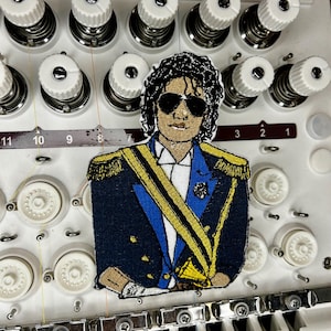 May include: An embroidered patch depicting a person with dark curly hair wearing black sunglasses and a blue military-style jacket with gold epaulets, a gold sash, and gold buttons, positioned on a white industrial embroidery machine.