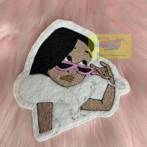 May include: Embroidered patch featuring a cartoon character with black hair, pink sunglasses, and a white dress. The patch is on a pink fuzzy background. The text "That's What She Threaded" is visible in the upper right corner.