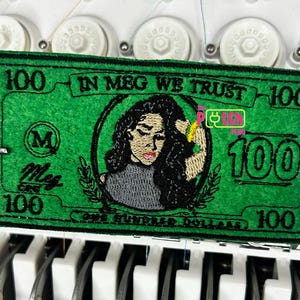 May include: Green embroidered patch resembling a $100 bill, featuring a portrait of a woman. The text "IN MEG WE TRUST" and "ONE HUNDRED DOLLARS" are visible. The patch is being worked on by an embroidery machine.