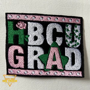 May include: An embroidered iron-on patch with the words "HBCU GRAD" in pink, green, and white letters on a black background. The patch has a pink border with white dots.
