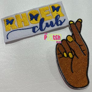 May include: Two embroidered iron-on patches. One patch is white with yellow text that reads "RHOER club" with blue butterfly accents. The other patch is brown with a yellow outline of a hand making a pinky promise.