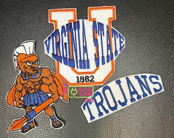 Può includere: Tre toppe ricamate con un emblema a forma di U bianco e arancione con il testo VIRGINIA STATE 1882 in blu, una figura di guerriero in armatura arancione e blu che impugna una lancia e una toppa bianca curva con il testo blu TROJANS.