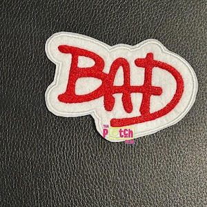 May include: An embroidered patch featuring the word BAD in bold red lettering with a white border. The letters have a stylized, slightly jagged appearance. A small logo with the text THA PATCH PLUG is visible at the bottom of the patch.