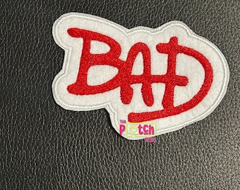 Può includere: Una toppa ricamata che presenta la parola BAD in lettere rosse audaci con un bordo bianco. Le lettere hanno un aspetto stilizzato e leggermente frastagliato. Un piccolo logo con il testo THA PATCH PLUG è visibile nella parte inferiore.