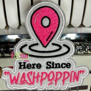 May include: Embroidered patch featuring a pink location pin design with a black outline, atop a white cloud-like shape. The text "Here Since WASHPOPPIN" is embroidered in black and pink. The patch has a gray border.