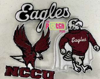 Può includere: Tre toppe ricamate. Una dice "Eagles" in corsivo bianco su sfondo nero. Un'altra mostra un'aquila marrone con le ali spiegate. La terza è una mascotte di aquila marrone che indossa un maglione con "Eagles". Sono su una superficie grigio chiaro.