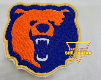 Può includere: Un toppa ricamata con la testa di un orso in arancione e blu, con una bocca e denti bianchi. La toppa ha un contorno dorato ed è su uno sfondo bianco. Il testo "That's What She Thread Embroidery" è visibile nell'angolo in basso a destra.